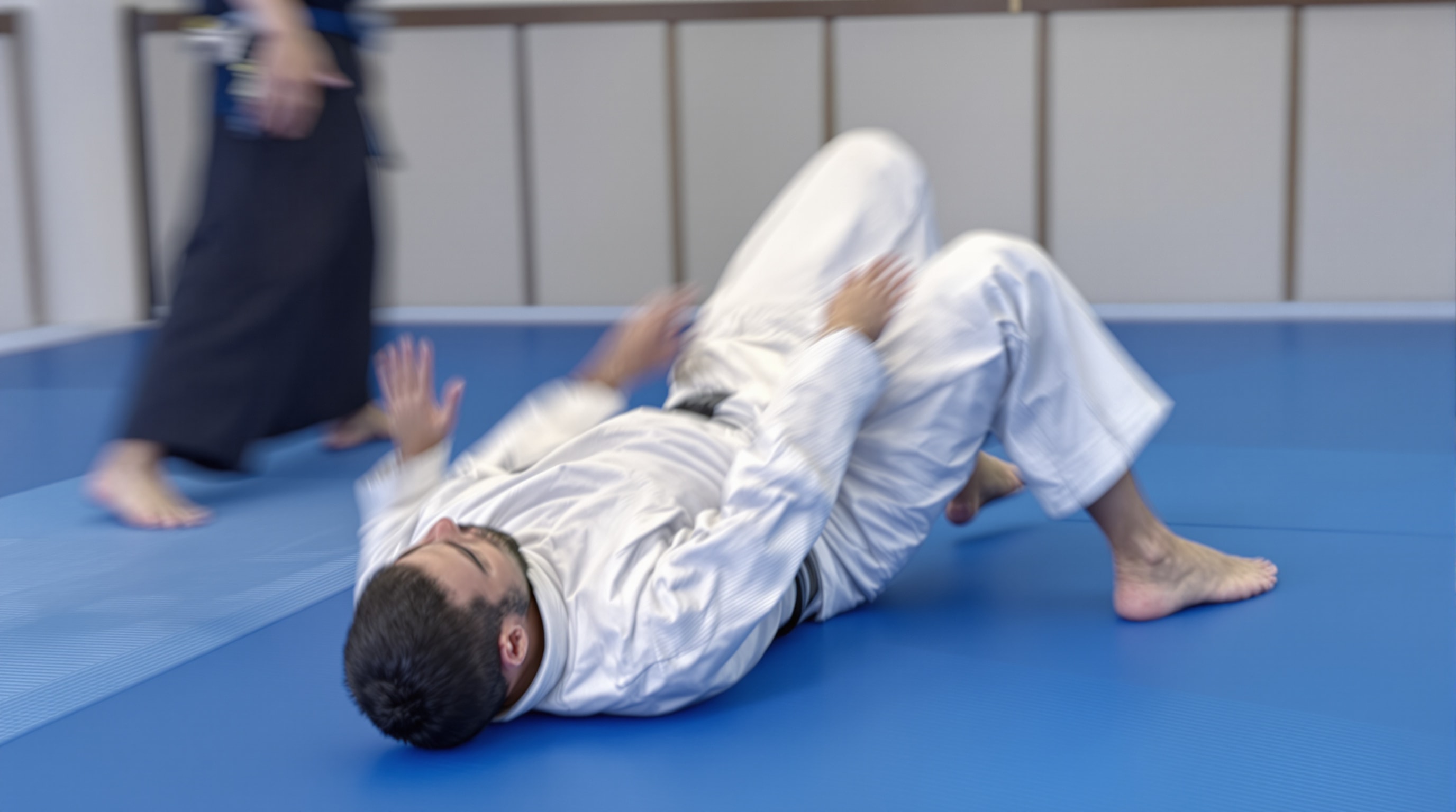 A beginner in a white gi practicing a forward rolling breakfall on blue tatami mats, seen from the side